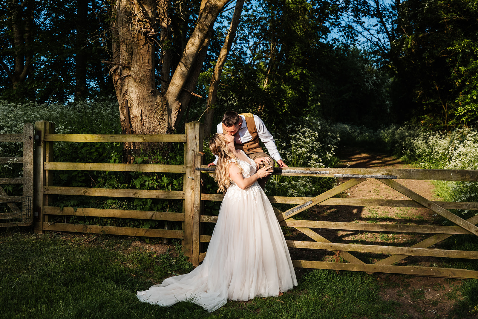 Couple share kiss with groom sitting on the top of a wooden gate, forest in the background 