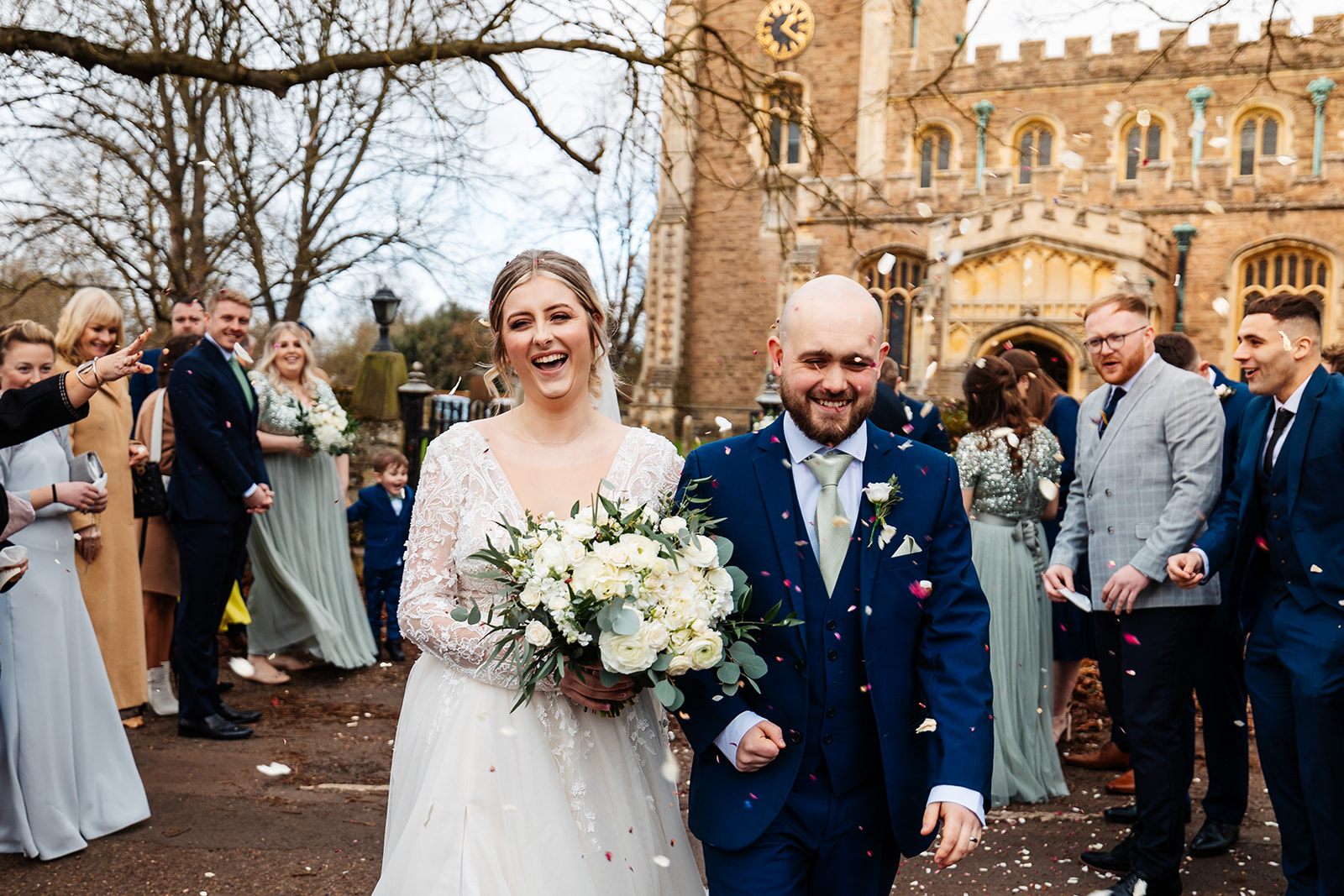 Couple walk away from church smiling whilst guests throw confetti 