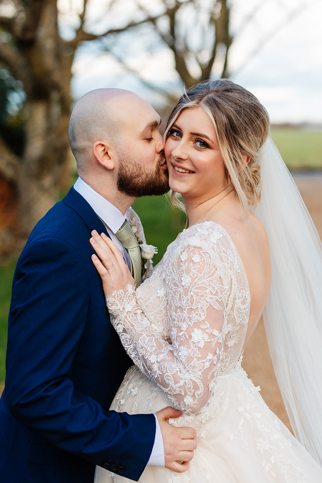 Bride looking straight down the camera whilst Groom kisses her on the cheek, tree lined driveway in the background