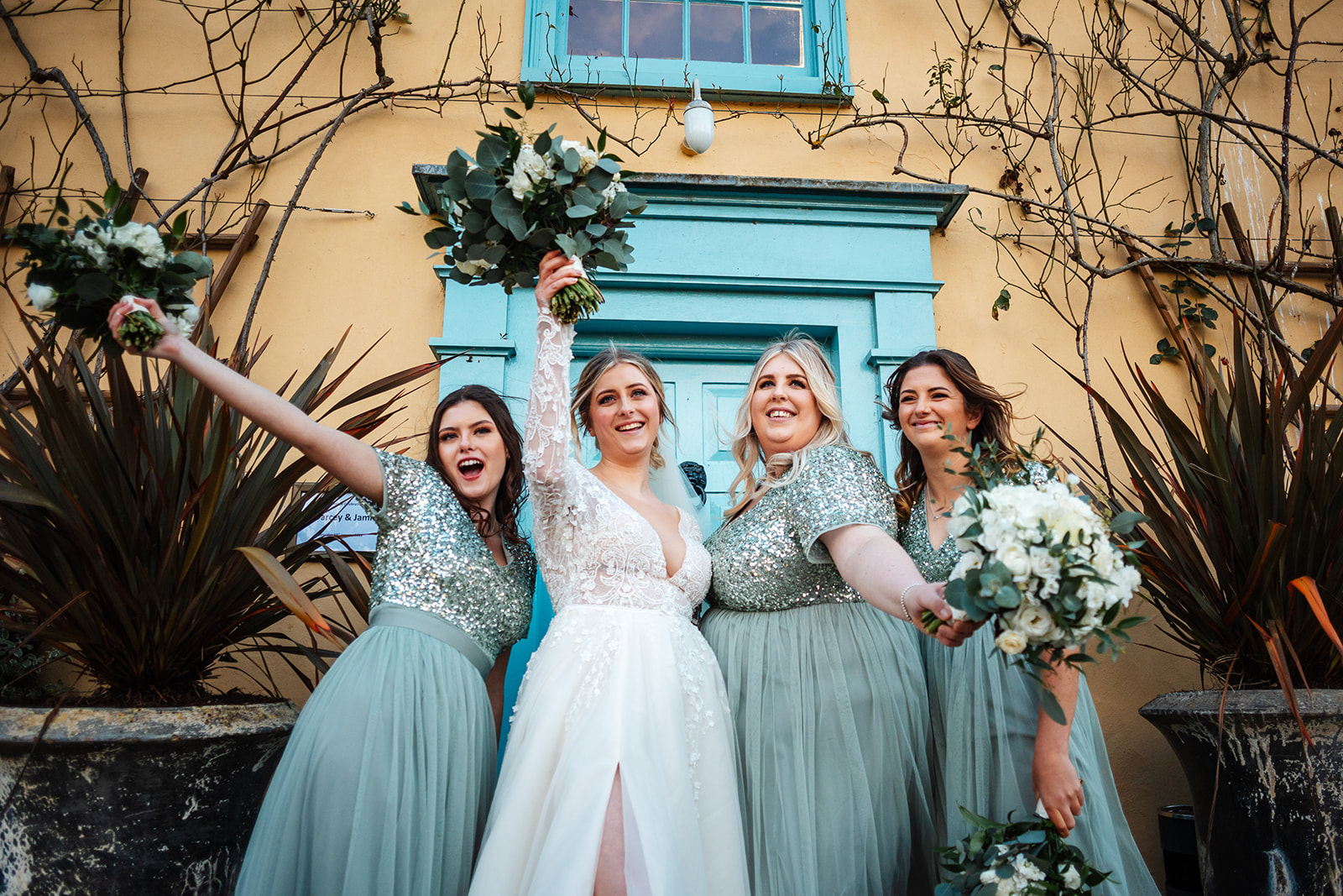 Bride and bridesmaids in green sequin dresses infront of main house at south farm, raising bouquets 