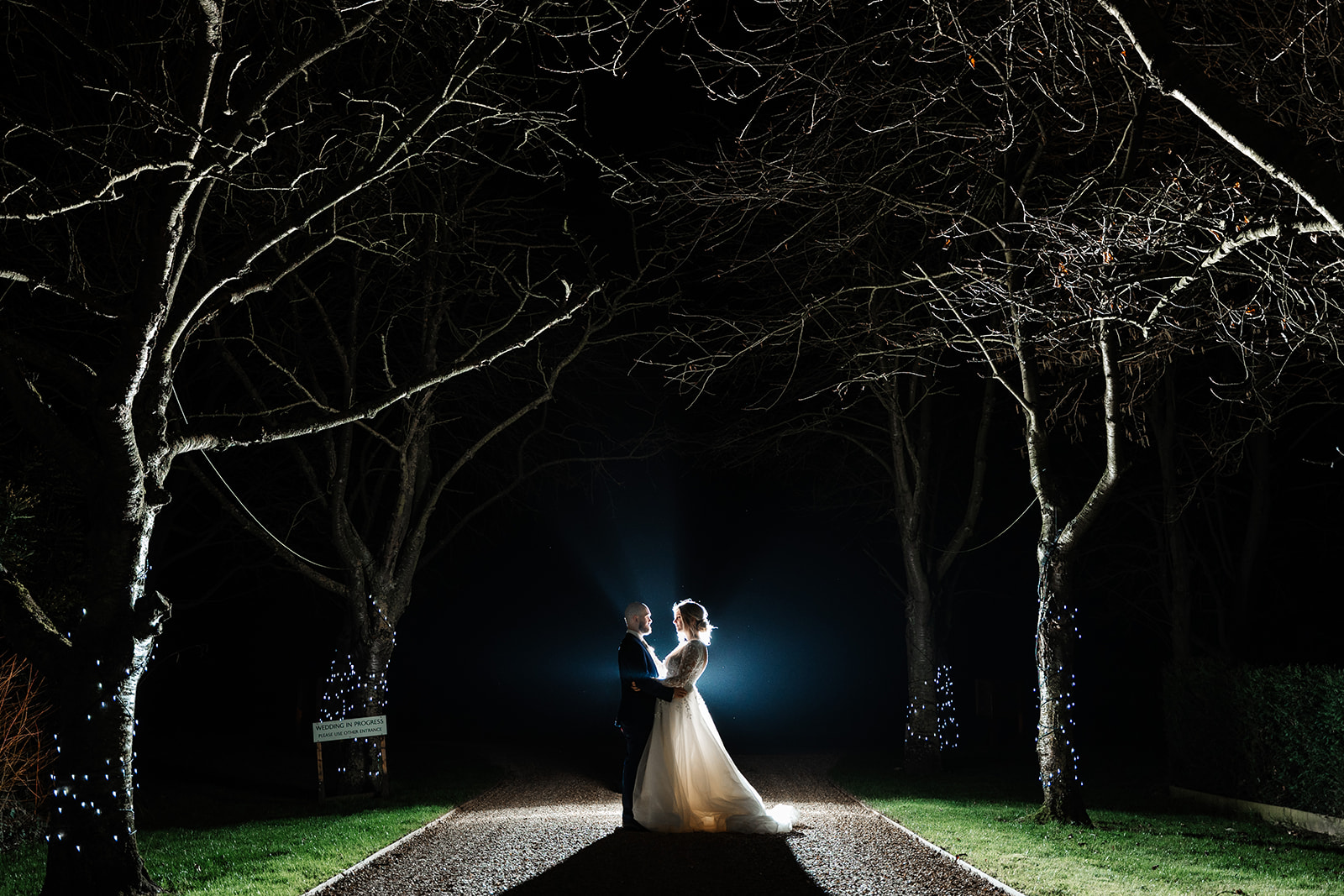 Couple on tree lined driveway at South Farm in the evening, fairy lights are on the trees 