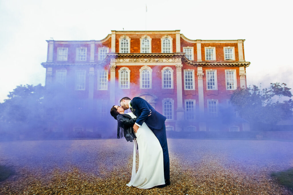 bride in leather jacket and boots and groom stood in front of chicheley hall surrounded by purple smoke