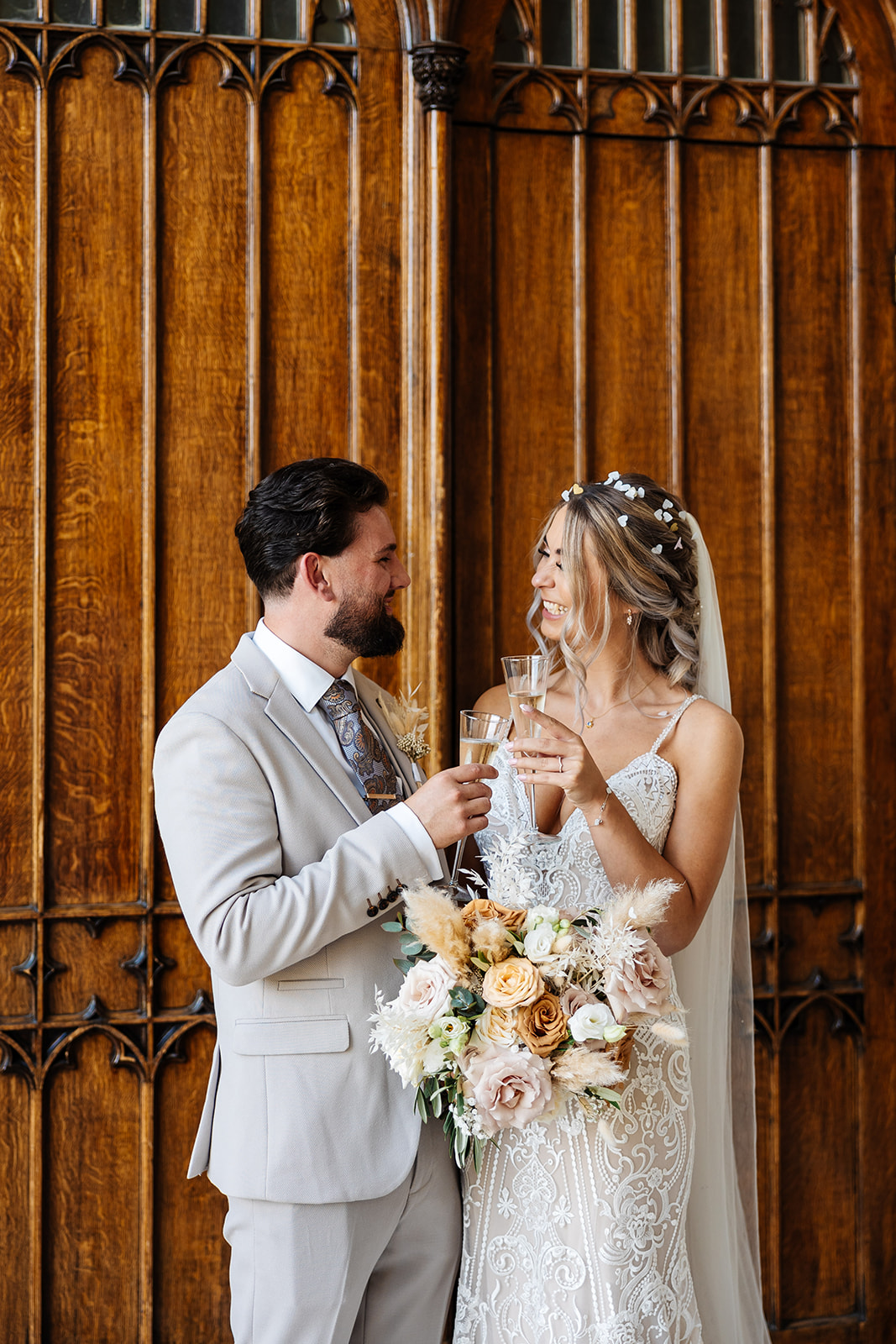 Bride & Groom toast to being married. They are stood infront of a wooden door inside Ashridge House. They are smiling at each other. 