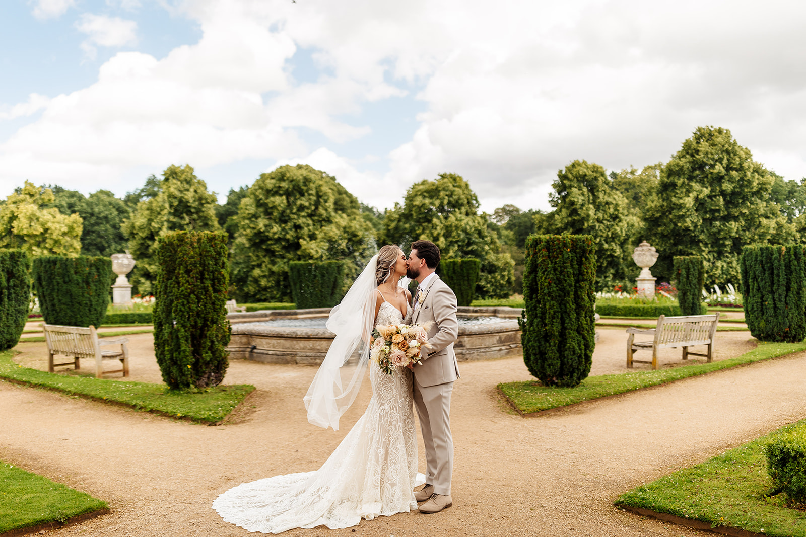 Bride & Groom share a kiss infront of the fountain, in the grounds of Ashridge House. Trees and gravel pathways are in the background. 