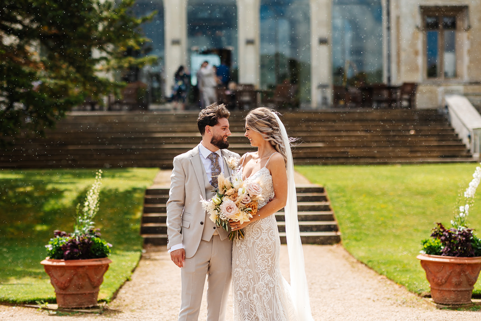 Couple outside Ashridge House, the steps to the house are behind them and they are stood on a gravel pathway, smiling at each other 