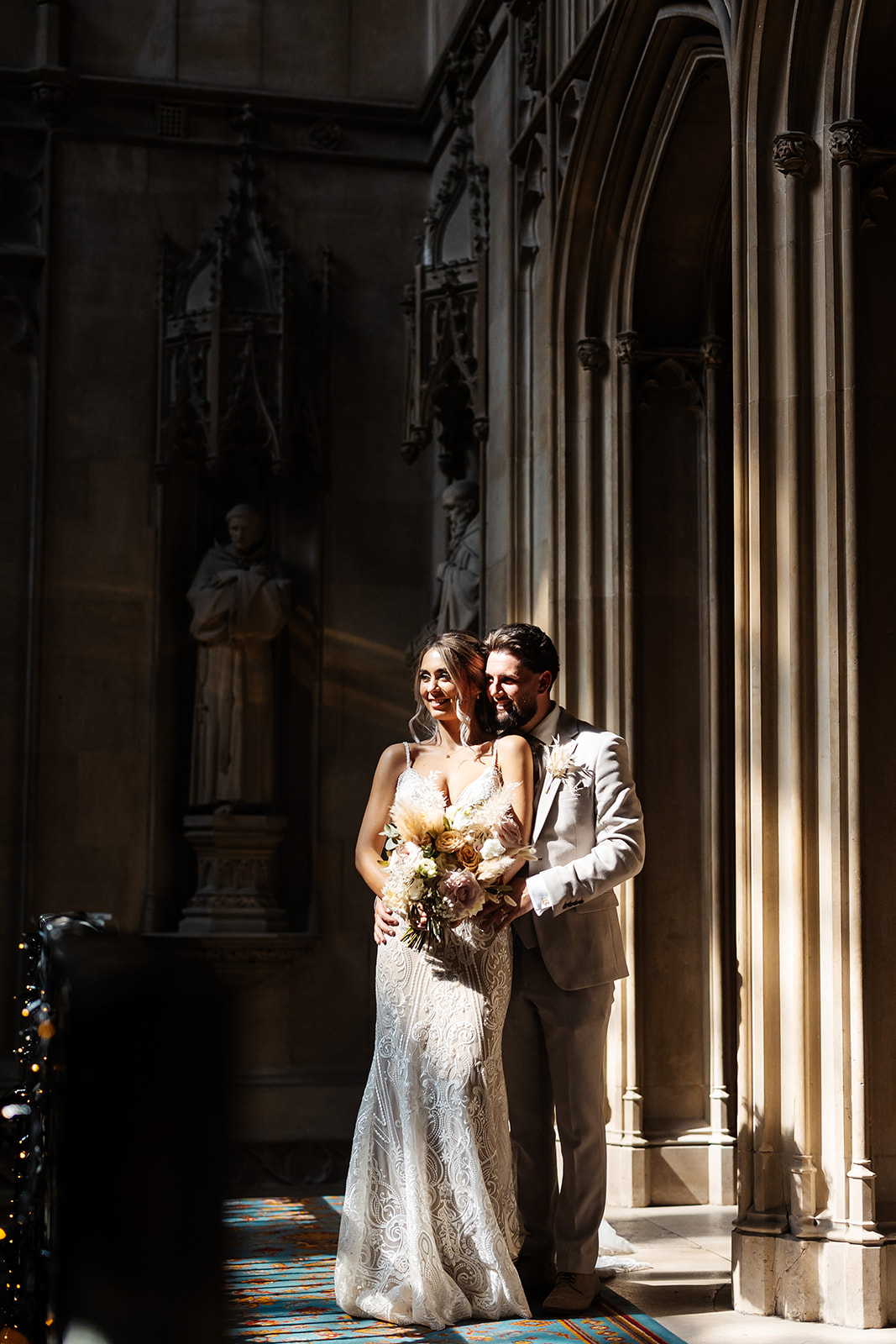 Bride and Groom on balcony inside Ashridge House, the light is hitting the stone pillars either side. The architecture of the interior is shown. 