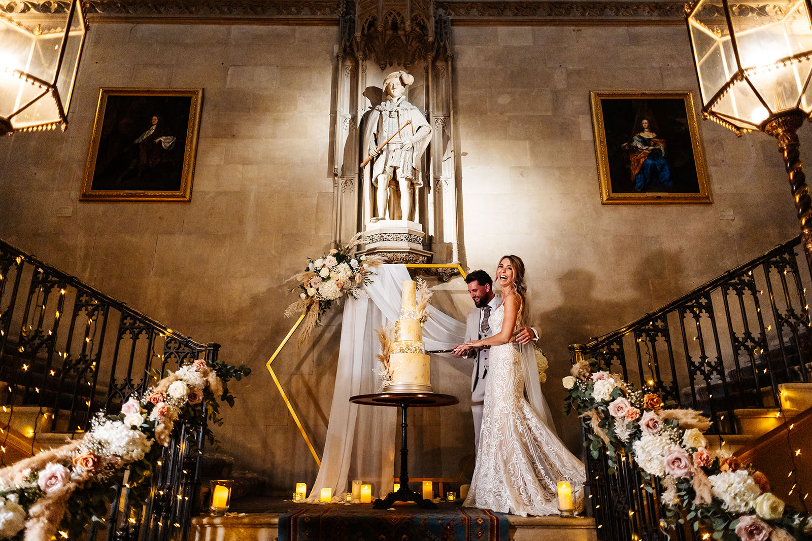 Couple cut their cake on the landing of the grand staircase inside Ashridge Manor. The cake is infront of floral backdrop, the bannisters are covered in florals and fairylights  and candles are lit on the floor 