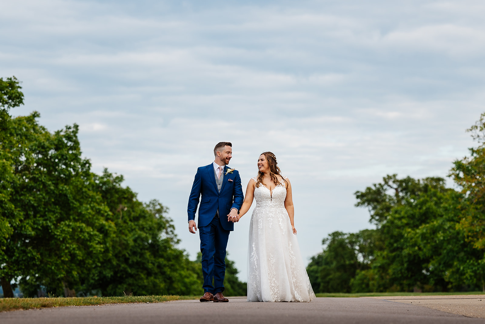 bride and groom walking hand in hand on driveway at bassmead manor barns, their are trees either side of them and a blue sky