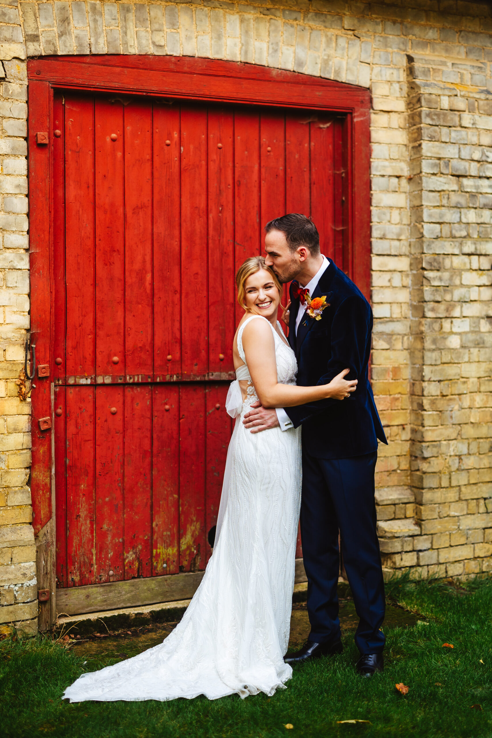 bride and groom stood close together in front of red barn door, groom is kissing bride on forehead at south farm 