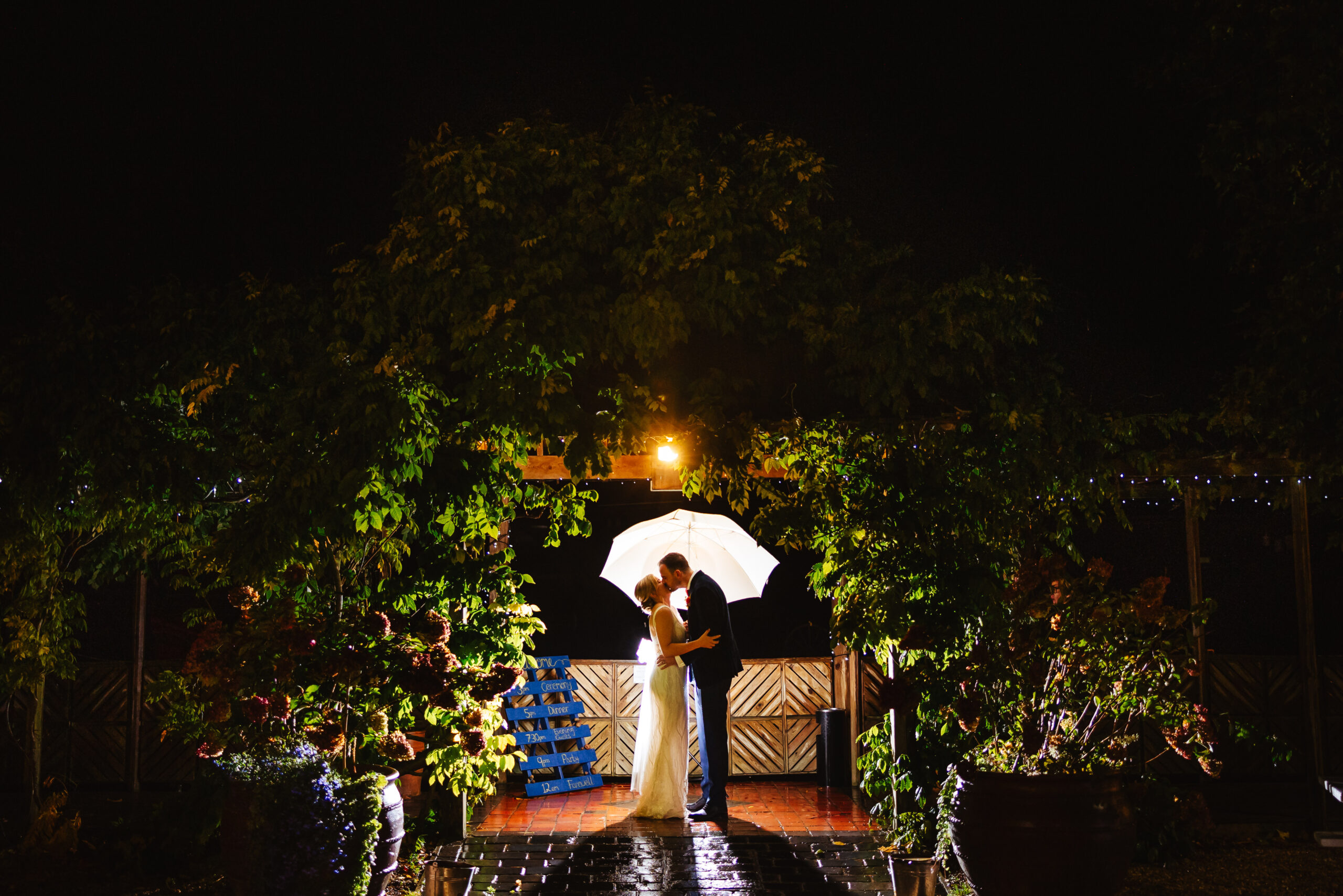 backlit barn entrance at south farm wedding, bride and groom have an umbrella and it is raining