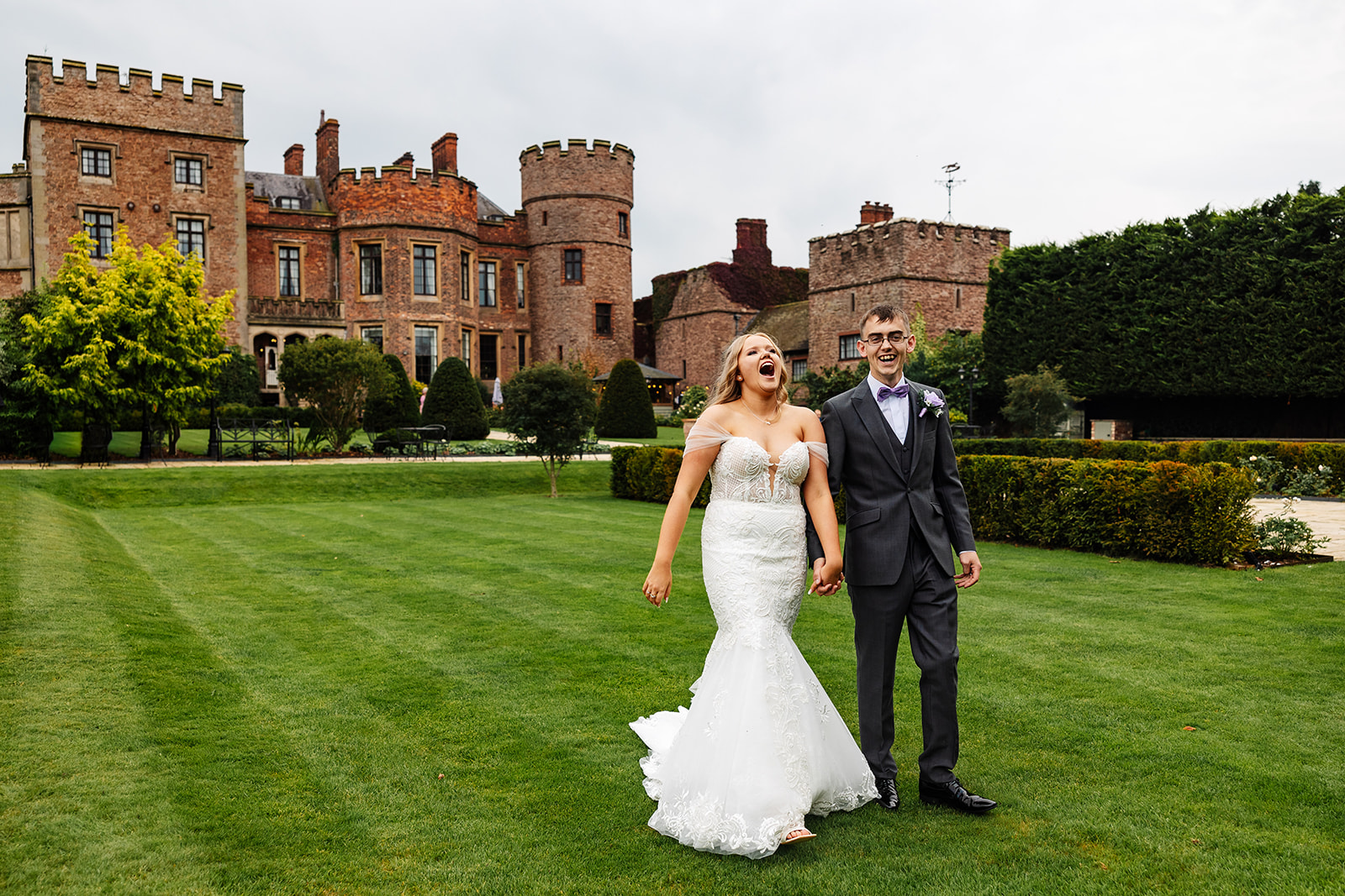 bride and groom stroll across green grass in front of rowton castle