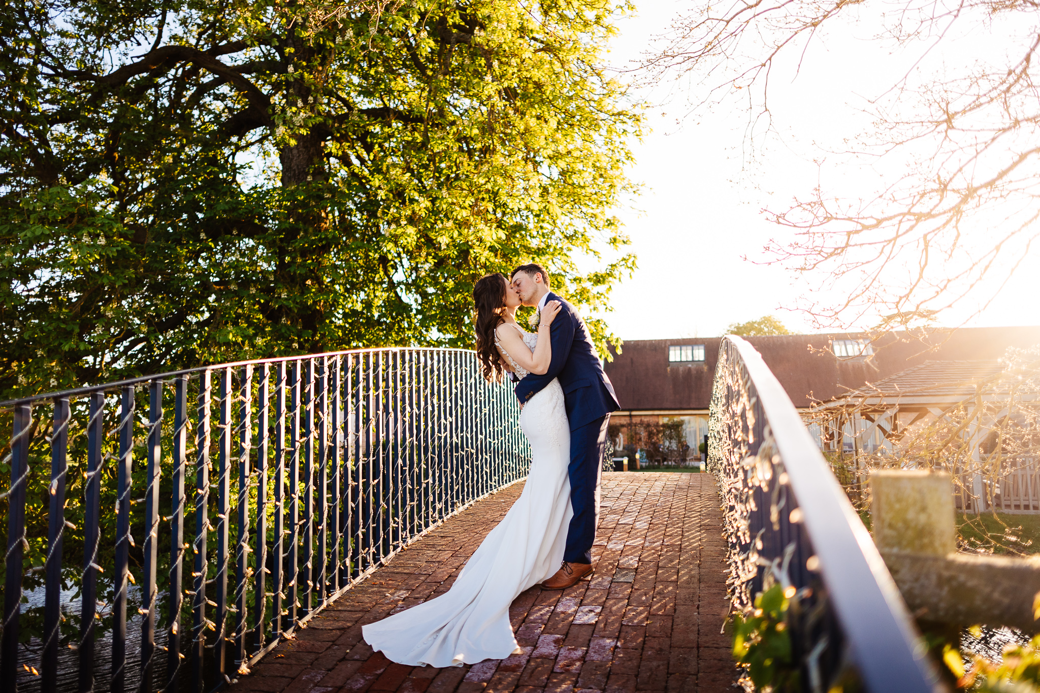 Bride and groom embracing on brick flooring blue bridge with fairy lights. The sun is shining through some trees as it goes down, at bassmead manor barns